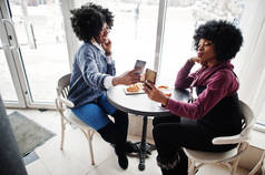 两个卷发非洲裔美国女人穿着毛衣坐在室内的咖啡馆里, 手里拿着电话.