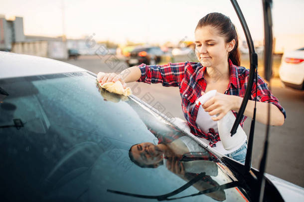 可爱的女人用海绵和喷雾, 洗车清洗汽车的前玻璃。女士在自助洗车。户外车辆清洁
