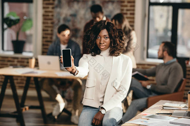 严重的非洲裔美国休闲女商人指着智能手机与空白屏幕在阁楼办公室与同事背后