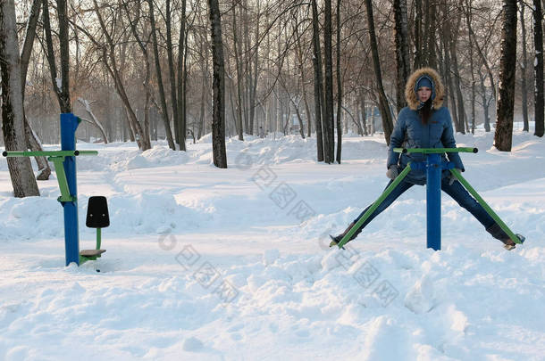 女人在蓝色<strong>羽绒服</strong>与胡德是做腿演习在模拟器在冬季公园前视图
