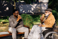 高级残疾人在轮椅和非洲裔美国人一起喝咖啡从纸杯一起在街上