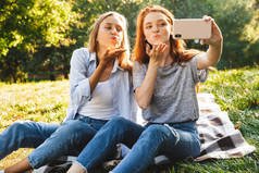 两个白人女孩的照片穿着休闲服装坐在草地上, 做空气亲吻, 而采取自拍智能手机在夏季公园