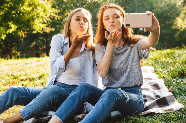 两个白人女孩的照片穿着休闲服装坐在草地上, 做空气亲吻, 而采取自拍智能手机在夏季公园