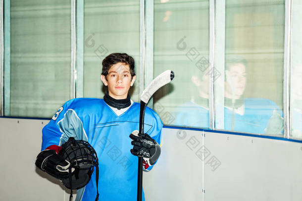 少年男孩的肖像, 职业冰球运动员在蓝色制服, 站立在溜冰场, 拿着头盔和棍子