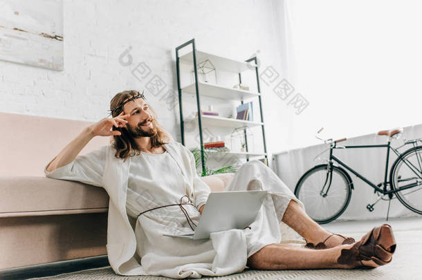 梦幻般的耶稣坐在地板上, 用笔记本电脑靠近沙发在家