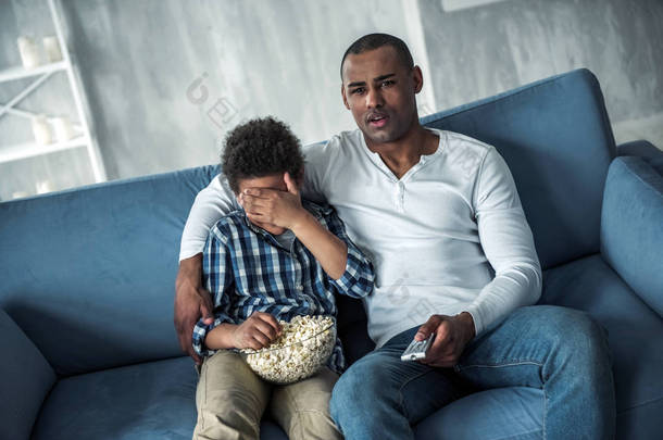 快乐的美国黑人父亲和儿子穿着便装在家里坐在沙发上看电视和吃爆米花。他们的面孔困惑, 男孩遮住了他的眼睛