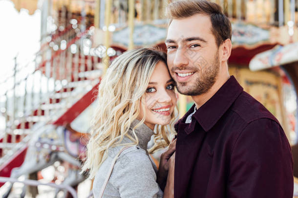 微笑深情情侣的肖像在秋季装备看摄像机附近的旋转木马在游乐园