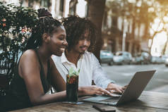 一个欢快的混合的情侣: 迷人的非洲女孩与辫子和英俊的亚洲年轻人卷曲的头发是通过笔记本电脑上网, 玩得开心, 街头咖啡馆, 饮料在桌子上