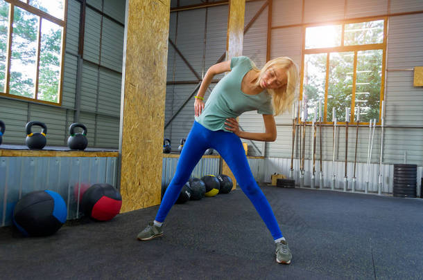在健身房的蓝色紧身裤的金发女郎是训练, <strong>运动器材</strong>背景倾斜斜坡: 医疗球, 哑铃和酒吧, 树木可以看到在窗口, 太阳的灯光