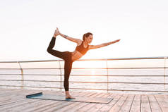 年轻的女士站在海边训练瑜伽。漂亮的女人在黑色运动的顶部和绑腿练习瑜伽与美丽的海景背景
