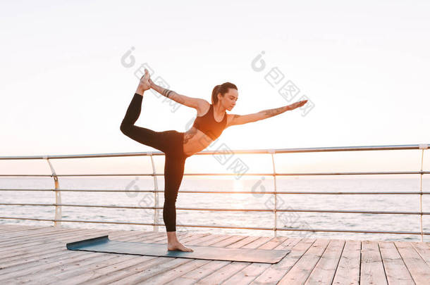 年轻的女士站在<strong>海边</strong>训练<strong>瑜伽</strong>。漂亮的女人在黑色运动的顶部和绑腿练习<strong>瑜伽</strong>与美丽的海景背景