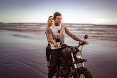 年轻时尚纹身情侣骑摩托车在海洋海滩上 