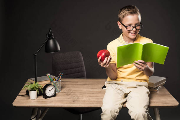 微笑的学校男孩与<strong>苹果</strong>做家庭作业和坐在桌上与书、灯、植物和颜色铅笔在<strong>灰色</strong>背景 