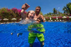 快乐活泼的家庭年轻父亲的有趣的照片活跃婴儿潜水在游泳池与乐趣跳跃在水下深处. 