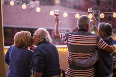四70岁的老人在阳台上庆祝用玻璃酒杯和红酒, 拥抱和亲吻对方。悬挂黄色灯泡, 创造气氛。幸福老人生活理念