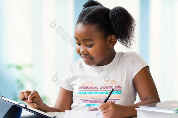 关闭可爱的小非洲女孩的肖像做家庭工作在办公桌上。Ponytailed <strong>儿童</strong>在数字平板电脑上打字和用钢笔<strong>写字</strong>.
