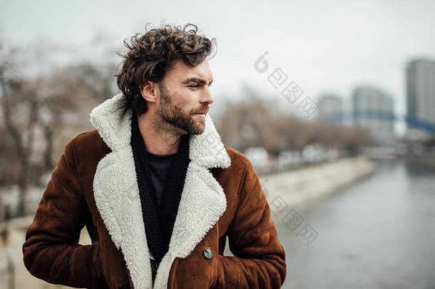 一个英俊的男人, 胡子, 冬天或秋季大衣站在外面的河边, 想某事或等待某人