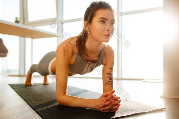 关闭在运动的顶部和绑腿漂亮的女士的照片练习健身瑜伽垫和听音乐在家里与大窗户的背景耳机