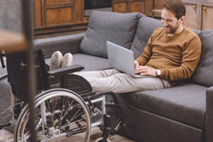 在家中使用膝上型电脑时, 微笑的残疾人把腿放在轮椅上