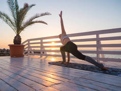 年轻的白种人女人做瑜伽体式之于自然木制阳台上俯瞰大海的桥。女人做了海洋在自然放松的练习。穿运动服的女孩.