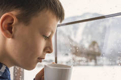 茶杯在手望着窗外没有在冬天雪山地森林景观的忧郁男孩