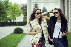 时尚拍摄的两个优雅美丽的女孩在夕阳的戴着墨镜，毛皮背心。两个年轻女人户外在大街上。购物的灵感