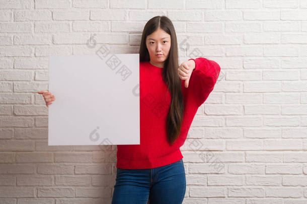 年轻的中国妇女在砖墙拿着横幅与愤怒的面孔, 消极标志显示不喜欢与拇指下来, 拒绝概念
