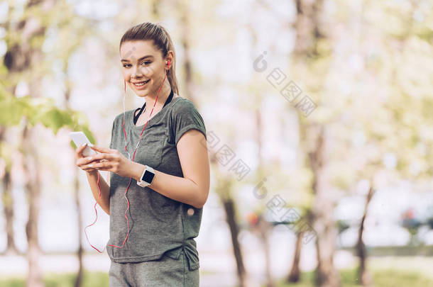 穿着运动服的漂亮女人在相机前微笑，同<strong>时</strong>拿着智能手机，在耳机里听音乐
