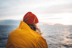 年轻的旅行者戴着红帽子和黄色的雨衣漂浮在船上，看着夕阳的大海，在暴风雨和雾山在天际线。生活方式旅行，斯堪的纳维亚地帽