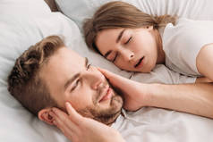 精疲力竭的男人靠在打鼾的妻子身边躺在床上，用手捂住耳朵