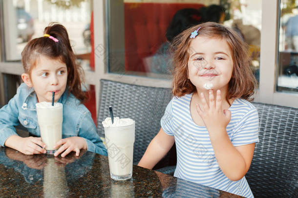 两个有趣的白人小妹妹在咖啡馆里喝着奶昔。朋友们一起玩的很开心给孩子们吃冷的夏季甜点。快乐而真实的童年生活. 