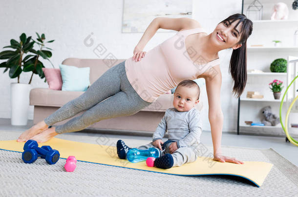 年轻快乐的母亲做侧板锻炼在瑜伽垫与她的孩子在家中