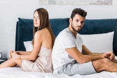 年轻的沮丧夫妇在睡衣坐在床上背靠背, 看着相机