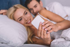 有选择的焦点, 年轻人偷看微笑的女朋友使用智能手机在床上, 不信任的概念