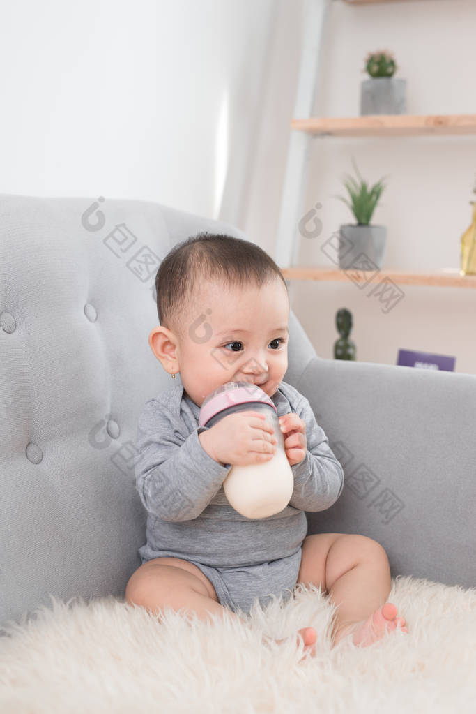 可爱的小女孩坐在沙发上, 从瓶子里喝着牛奶, 微笑着。快乐的婴儿。家庭人室内的内部概念。童年的最佳时间!