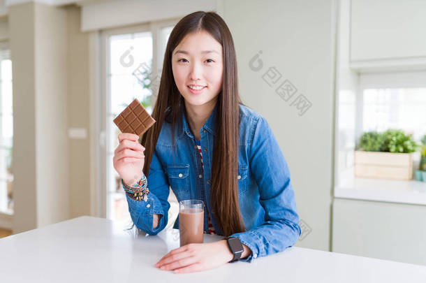 美丽的亚洲女人喝巧克力奶昔，拿着巧克力棒，一张幸福的脸站着微笑，带着自信的微笑露出牙齿