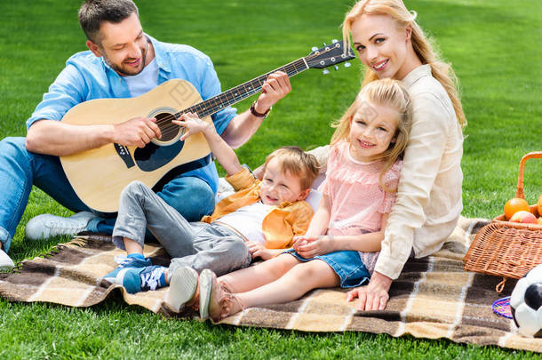 快乐的家庭玩吉他和坐在一起在格子在野餐 