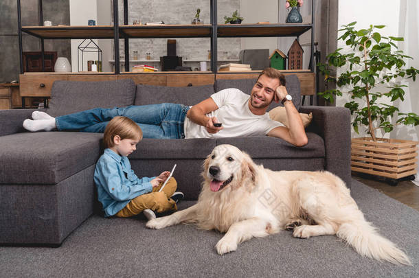 快乐的父亲看电视在沙发上, 而他的儿子用平板电脑在<strong>地板</strong>上与金猎犬