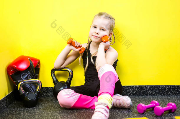 主题体育和健康儿童。小搞笑的孩子白种女孩与辫子，坐在<strong>健身房</strong>的地板上<strong>休息休息</strong>。运动员哑铃器械为体操健美背景黄墙