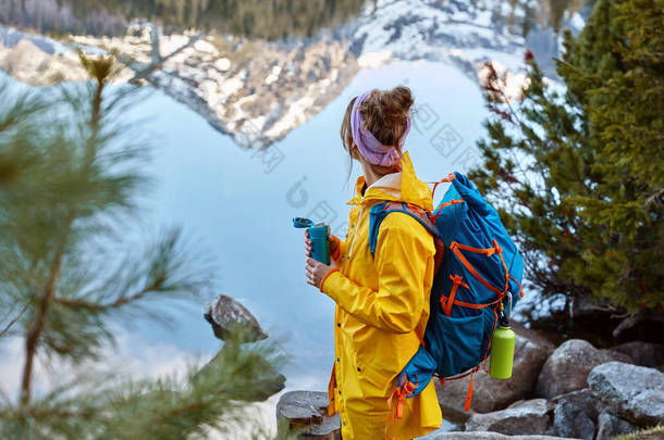 这位徒步旅行的妇女在山上的湖边停下来，背着背包，拿着<strong>热饮料</strong>的保温瓶，探险