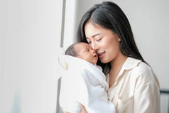 白衬衫亚洲母亲亲吻她的新生儿在卧室前的玻璃窗与白色窗帘，以显示爱和家庭纽带.