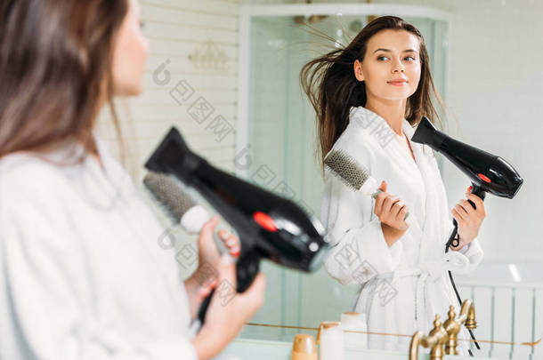 美丽的年轻妇女举行头发刷子和干燥头发在<strong>镜子</strong>在浴室