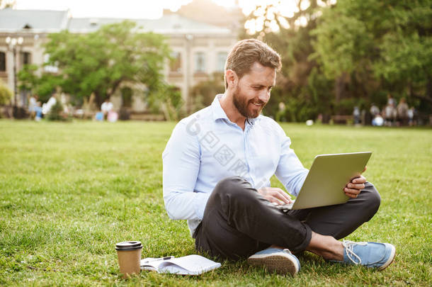 商业服装的务实高兴的人的照片坐在草地上, 腿交叉和工作在银色笔记本电脑
