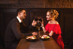 年轻的情侣在餐厅, 浪漫的约会。典雅的妇女在红色礼服和英俊的人餐饮和饮用的酒