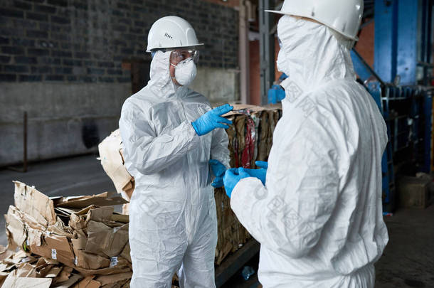 两名身穿生物防护服的工人画像现代废物处理厂工业仓库中的通讯, 背景下的可回收纸板堆