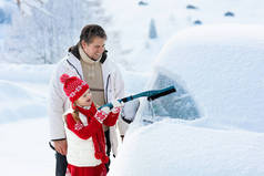 父亲和孩子在暴风雨后刷洗和铲去汽车上的雪.带着冬刷和刮胡刀的父母和孩子在一夜暴风雪之后清扫了家里的汽车。山里的圣诞假期.