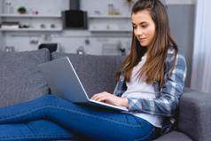 微笑的年轻女子坐在沙发上, 使用笔记本电脑 