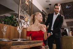 英俊的男朋友帮助坐下来有吸引力的女朋友在红色的礼服, 微笑着, 拿着椅子