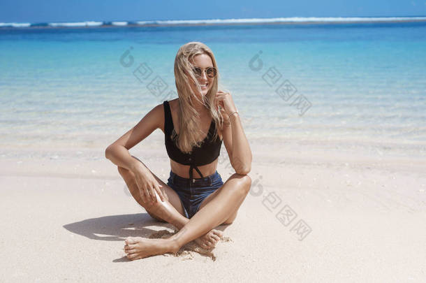旅游，旅游，度假理念。有吸引力的快乐放松晒<strong>黑金</strong>发欧洲妇女坐在沙滩附近的海岸面对相机微笑快乐日光浴，享受暑假
