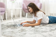 迷人的年轻黑发女人穿着牛仔裤和白色T恤躺在明亮的客厅与她的笔记本电脑和智能手机的地板上